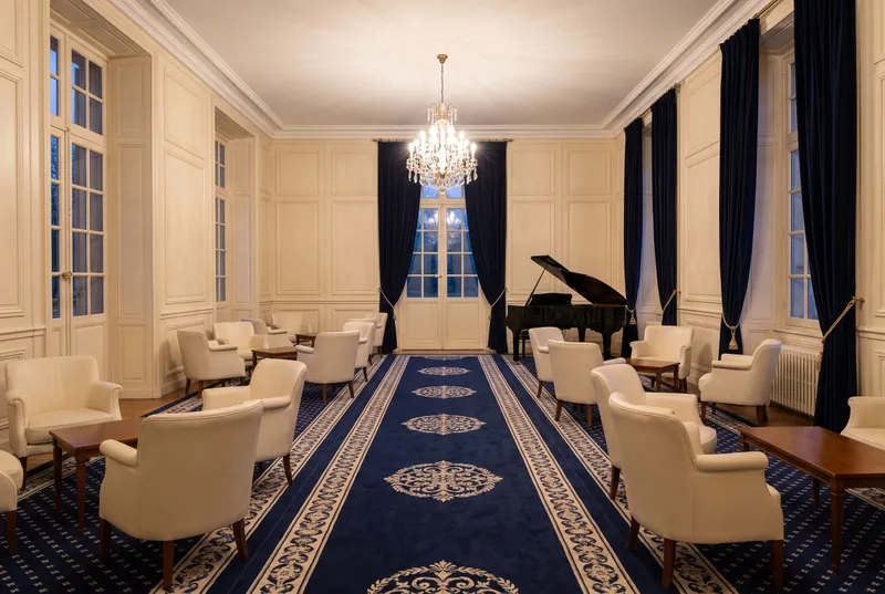 Scène générée : un grand hall lambrissé de crème, un long tapis bleu marine, des fauteuils, un piano à queue fermé et un lustre allumé.