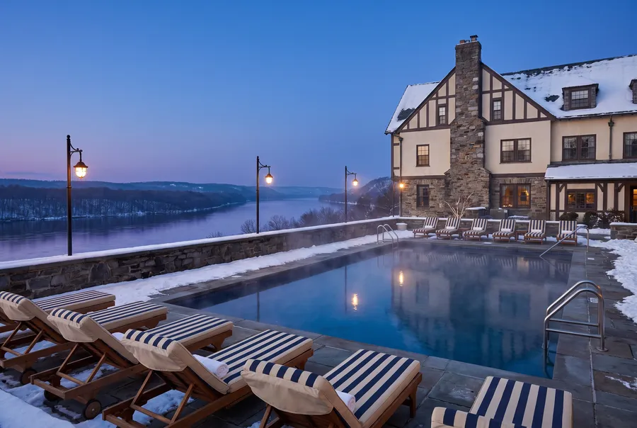 Scène générée : une piscine extérieure chauffée en hiver au crépuscule, de la vapeur sur l'eau, de la neige sur la terrasse et un fleuve en contrebas.